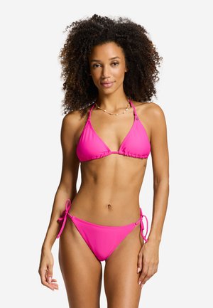 Kvinde med krøllet hår iført en lyserød snøre-bikini, der står foran en ensfarvet hvid baggrund, vendt fremad med et let smil.