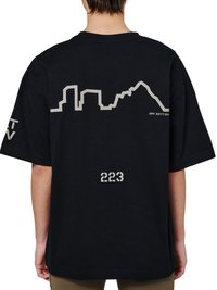 Personne portant un T-shirt noir oversize avec un motif de silhouette de ville grise et le texte "OFF DUTY GEAR" sur le haut du dos, et "223" près du bas du dos.