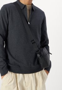 Svart crossover-väska med dragkedjefack, slät textur, fäst vid ett långt justerbart band, bärs över en mörkgrå tröja och krämfärgad byxor.