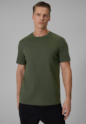 Mężczyzna w prostym oliwkowozielonym t-shircie z krótkim rękawem i czarnych spodniach stoi na tle jasnoszarym.