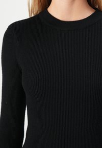 Detalhe de uma pessoa a usar um suéter preto de malha canelada com gola redonda e mangas compridas.
