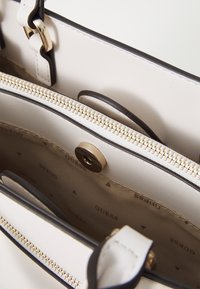 Una borsa a tracolla di colore crema con dettagli neri, dotata di una cerniera dorata, chiusura a pulsante e un interno decorato con il nome del marchio ripetuto.