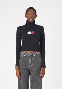 Maglione a collo alto nero a coste con logo ricamato "TOMMY JEANS" sul petto, abbinato a jeans grigi in denim a vita alta.