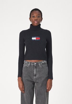 Maglione a collo alto nero a coste con logo ricamato "TOMMY JEANS" sul petto, abbinato a jeans grigi in denim a vita alta.
