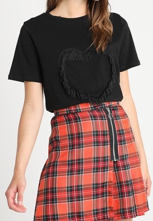 Kvinde iført sort t-shirt med frynset hjerte design og orange-rødt ternet nederdel med lynlås foran.