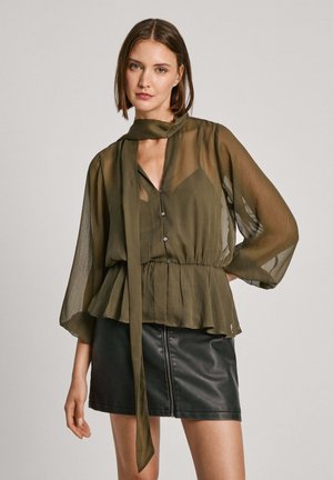 Blouse olive vert, translucide, à manches longues avec une taille peplum et des détails de boutons sur la poitrine. Associée à une mini-jupe en cuir noir.