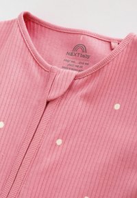 Närbild av rosa ribbad babyplagg med vita prickar och tryckknappsstängning, visar NEXT baby-märke och rund halsringning.