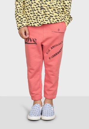 Koralle Jogginghosen mit schwarzem Drucktext. Kombiniert mit einer gelben gemusterten Jacke und blauen Slippern mit weißen Sohlen.