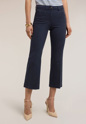 Vrouw met marineblauwe, wijd uitlopende, cropped broek, gestreept shirt, beige hoge hakken en gouden armbanden om haar linkerpols, staand tegen een lichte achtergrond.