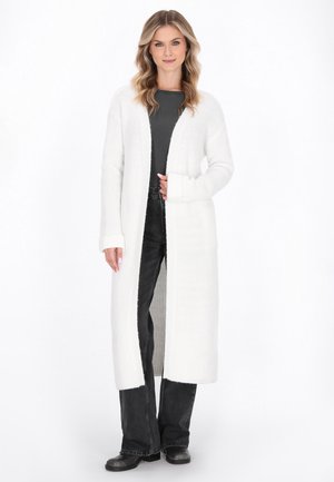 Nainen, jolla on pitkä valkoinen neulecardigan mustan paidan, mustien leveälahkeisten farkkujen ja mustien saappaiden päällä, seisoo valkoista taustaa vasten.