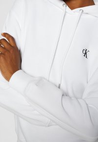 Personne à la peau brune moyenne croise les bras, portant un hoodie blanc Calvin Klein avec logo cK visible et cordons de serrage.