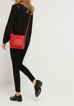 Rød læder crossbody taske med en enkelt skulderrem og lomme med lynlås foran. Båret med sort top og tætsiddende sorte bukser. Sorte sko.
