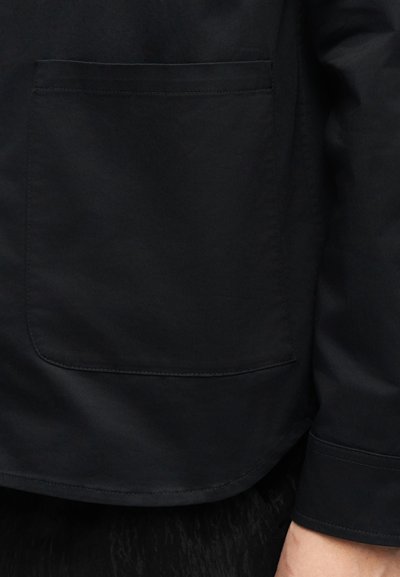 Μαύρο μακρυμάνικο πουκάμισο με μεγάλη τσέπη μπροστά και εμφανή ραφές κοντά στο τελείωμα και το μανίκι, φορεμένο από ένα άτομο.