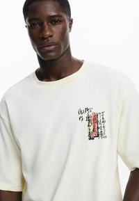 Camiseta de algodón blanca con un diseño impreso en negro y rojo que incluye texto abstracto. La camiseta tiene un corte holgado y un cuello redondo.