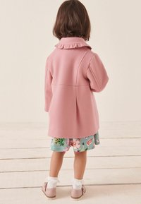 Light pink wool coat with ruffled collar and long sleeves, paired with floral print shorts. White frilly socks and pink shoes complete the outfit.