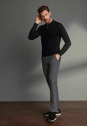 Schwarzes Langarm-Polohemd, graue maßgeschneiderte Hose und schwarze Schuhe mit weißen Sohlen, stehend auf einem Holzfußboden vor einem grauen Hintergrund.