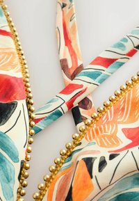 Audrinis audinio vaizdas su spalvingu abstrakčiu lapų raštu ir auksinių karoliukų apdaila ant susikertančių dirželių.