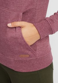 Burgundivörös kapucnis pulóver, puha anyagból készült, kangouru zsebbe, bordás mandzsettával és egy kis bőr logó címkével az elején.
