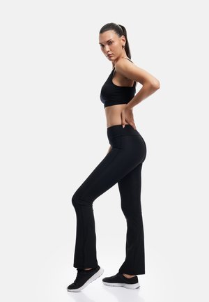 Kobieta w czarnym sportowym biustonoszu i legginsach stoi w pozycji bocznej z jedną ręką na dolnej części pleców i czarnych trampkach na białym tle.