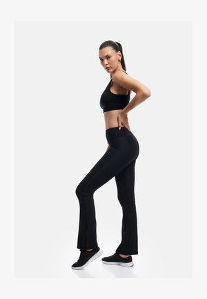 Kvinde i sort sportstop og leggings står i sidepose med den ene hånd på lænden og sorte sneakers mod hvid baggrund.
