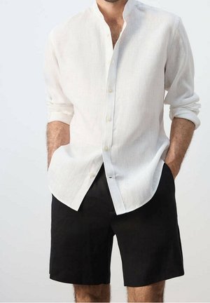 Homme portant une chemise blanche à manches longues retroussées et un short noir jusqu'aux genoux, les mains dans les poches.