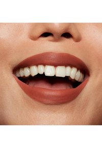 Närbild av en leende mun med matt terrakottaläppstift, vita tänder och en slät hudtextur. Naturlig läppform och subtila skuggor syns.