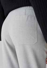 Graue Hose aus strukturiertem Stoff, mit einer einzelnen Gesäßtasche und geradem Schnitt. Ein schwarzer Strickpullover ist teilweise sichtbar.