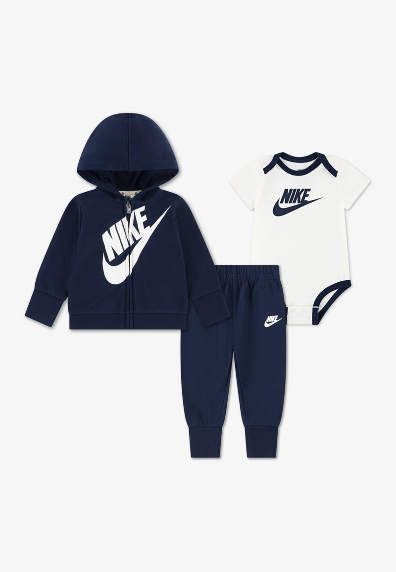 Σετ βρεφικής φόρμας σε ναυτικό μπλε που περιλαμβάνει φούτερ με φερμουάρ και μεγάλο λευκό λογότυπο Nike, ταιριαστό παντελόνι και λευκό φορμάκι με ναυτικά στοιχεία.