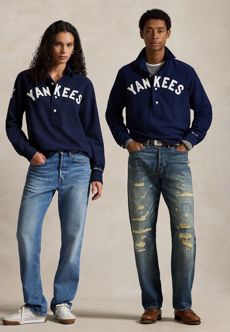 Camisas de manga longa azuis da Marinha com a inscrição "YANKEES", combinadas com jeans claros e desgastados; ténis brancos em um, sapatos castanhos no outro.
