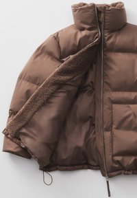 Blouson rembourré marron avec une fermeture éclair à l'avant, présentant une doublure en sherpa texturée le long du col et des poches, avec un extérieur lisse et brillant.