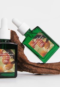 Two green dropper bottles of gentle vitamin C serum with 20% fruit water, placed on and beside a textured piece of wood.