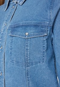 Cămașă din denim de culoare albastru deschis, cu guler, butoane tip capse și două buzunare frontale cu clapete. Detalii vizibile de textură și cusături sunt prezente.