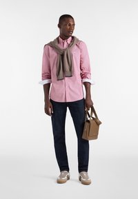 Camicia button-up rosa chiaro, maglione beige annodato intorno al collo, jeans scuri e sneakers beige, mentre tiene una borsa marrone con un logo.