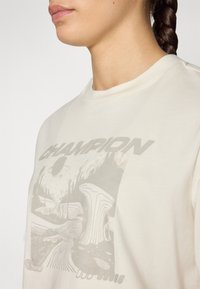 Helles beiges Sweatshirt mit Rundhalsausschnitt, ausgestattet mit einem grauen grafischen Design einer Landschaft und dem Text "CHAMPION" in fetten Buchstaben.