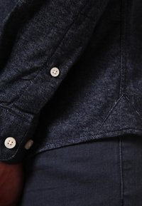 Donkerdenim overhemd met een gestructureerde stof, voorzien van witte knopen en een net gestikte kraag. Close-upweergave toont de details van de zoom en de manchetten van het overhemd.