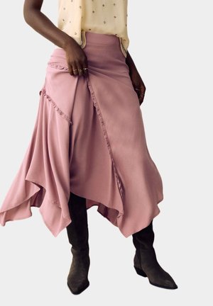 Mujer con una falda asimétrica rosa polvoriento fluida con ribete de volantes y botas negras de punta puntiaguda, sosteniendo la tela de la falda con la mano.