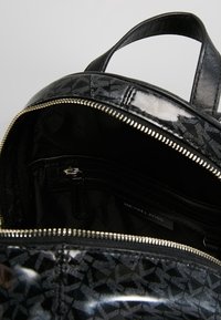 Zaino nero realizzato in materiale lucido con motivo a rilievo. L'interno presenta una tasca con zip e un'etichetta di branding. Chiusura con zip dorata.