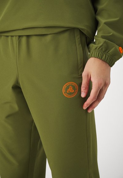 Olivenfarbene Sporthosen mit glattem Material, elastischem Bund und einem kleinen orangen runden Logo auf dem rechten Oberschenkel.