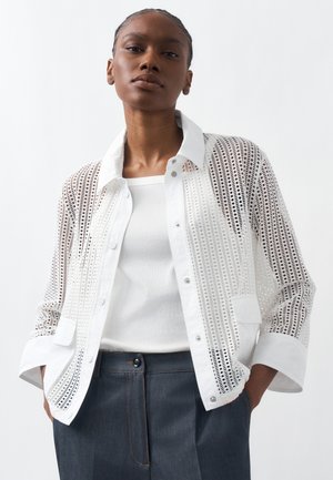 Woman wearing a white perforated button-up shirt over a white tank top, paired with dark high-waisted pants, standing with hands in pockets.