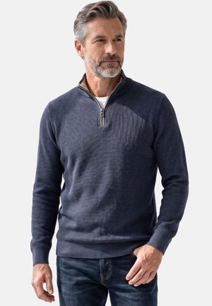 Mann mit grauem Haar und Bart trägt einen marineblauen, strukturierten Pullover mit Viertel-Reißverschluss und dunkle Jeans, blickt seitlich mit neutralem Ausdruck.