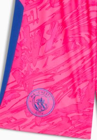 Jasnoróżowy sportowy trykot z teksturowanym graficznym wzorem, z niebieskimi akcentami i okrągłym logo Manchester City na dole.