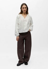 Femme portant un chemisier blanc à manches longues à motifs floraux et un pantalon large foncé, debout avec une main dans la poche, sur fond blanc.