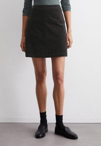 Zwarte denim rok met een getailleerde pasvorm, knie-lang, met een voornaad en een eenvoudige zoom. Gecombineerd met zwarte instappers en donkere sokken.