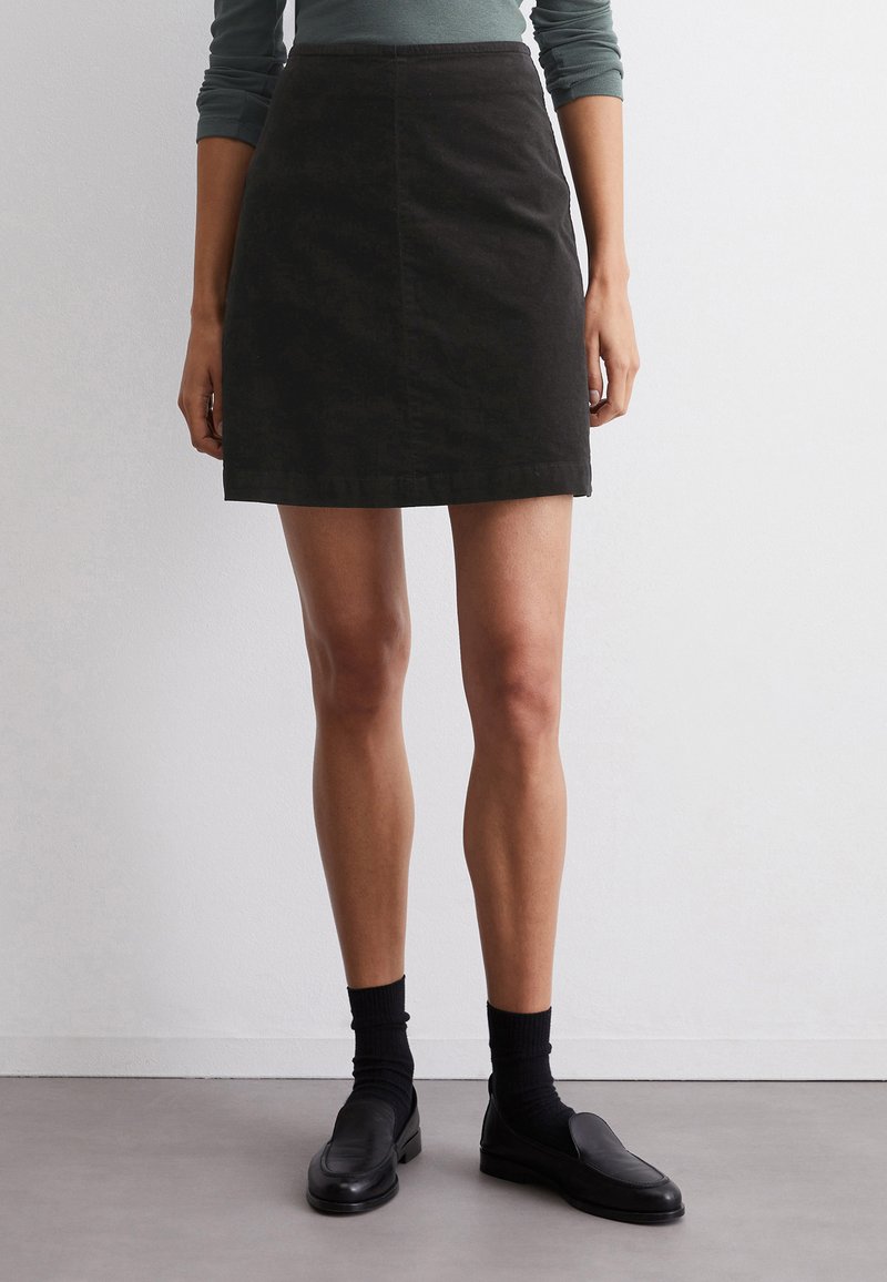 Zwarte denim rok met een getailleerde pasvorm, knie-lang, met een voornaad en een eenvoudige zoom. Gecombineerd met zwarte instappers en donkere sokken.