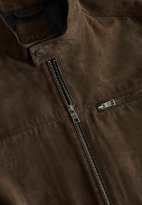 Rudos spalvos zomšinė striukė su priekyje metaline užtrauktuku, užtrauktuku ant krūtinės kišenės ir saga susegamu apykakle, viduje pažymėta "NEXT".