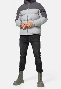 Veste matelassée grise et noire avec capuche, dotée de poignets élastiques et d'une fermeture éclair, portée avec un jean foncé et des bottes grises à fermeture éclair.