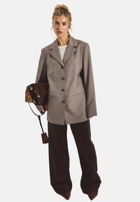 Übergroßer grauer Blazer aus strukturiertem Stoff, mit auffälligen Knöpfen und einem Reverskragen. Kombiniert mit dunklen, weit geschnittenen Hosen und einer glänzenden Handtasche.