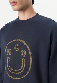 Sudadera azul marino con un diseño de cara sonriente bordado en hilo dorado en forma circular, con el texto "HGO" integrado en el patrón. Material de mezcla de algodón.