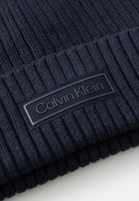 Navy knitted beanie featuring ribbed texture and a black rectangular tag with "Calvin Klein" embossed. Soft material, snug fit.