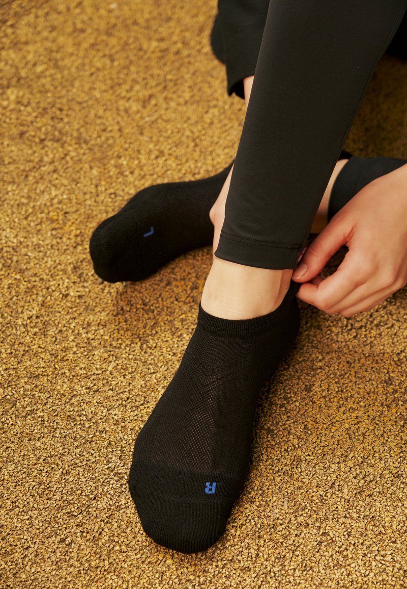 Person, die schwarze Knöchelsocken mit blauen linken und rechten Buchstaben trägt, passt einen Socken an, während sie auf einem strukturierten braunen Boden sitzt.
